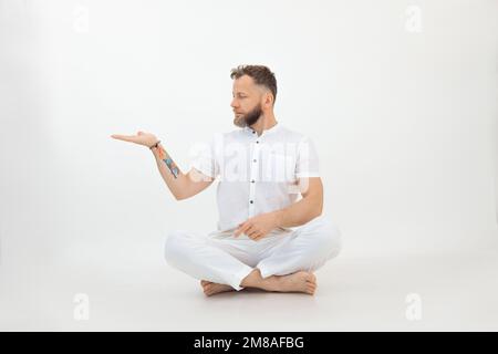 Ein Mann mittleren Alters mit Bart sitzt mit gekreuzten Beinen auf dem Boden und hebt die Hand mit offener Handfläche auf weißem Hintergrund. Stockfoto