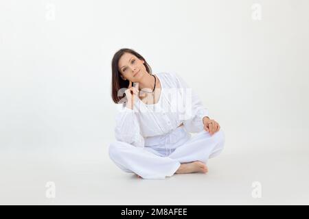 Porträt einer jungen Frau mit kurzen dunklen Haaren in weißer, lockerer Bluse, Hose, die mit gekreuzten Beinen auf dem Boden sitzt. Stockfoto