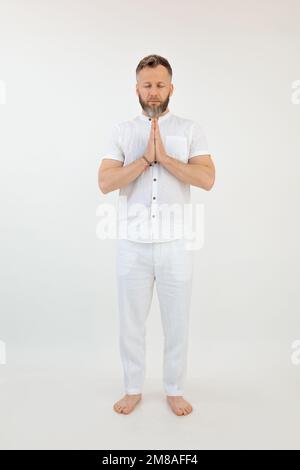 Konzentrierter, sportlicher bärtiger Mann mittleren Alters mit geschlossenen Augen im Stehen, faltbare Hände auf weißem Hintergrund. Meditation. Stockfoto