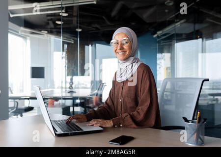 Porträt einer erfolgreichen Geschäftsfrau im Büro mit Laptop, einer Frau im Hidschab, die lächelt und in die Kamera schaut, einer muslimischen Büroangestellten, die eine Brille trägt. Stockfoto