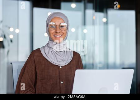 Porträt einer erfolgreichen Geschäftsfrau im Büro mit Laptop, einer Frau im Hidschab, die lächelt und in die Kamera schaut, einer muslimischen Büroangestellten, die eine Brille trägt. Stockfoto