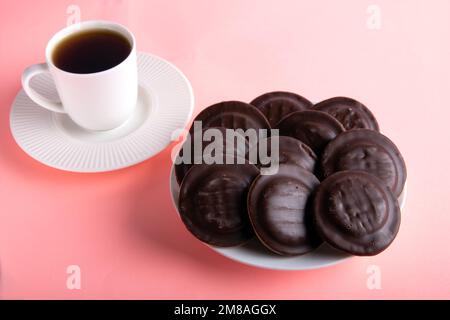 Runde Schokoladenkekse auf einem weißen runden Teller neben einer weißen Tasse heißes Getränk Stockfoto