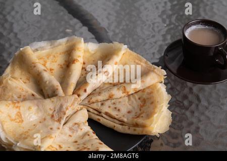 Machen Sie Fotos von dreieckigen gebratenen Pfannkuchen, die in einem Kreis auf einem Teller gestapelt sind, und einer dunklen Tasse Kaffee auf einem Glastisch Stockfoto