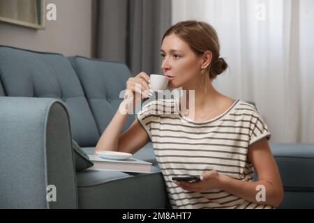 Entspannte junge Frau, die eine Tasse Kaffee im Wohnzimmer genießt, entspannender Selbstpflegetag zu Hause Konzept. Stockfoto