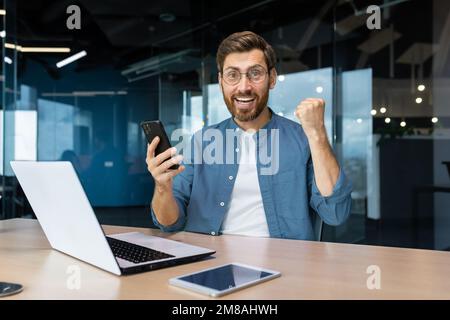 Porträt eines Geschäftsmanns, der mit den Ergebnissen in einem modernen Büro zufrieden ist, ein Mann feiert einen Sieg, schaut auf die Kamera hält ein Telefon in der Hand, ein Programmierer verwendet ein Notebook bei der Arbeit. Stockfoto