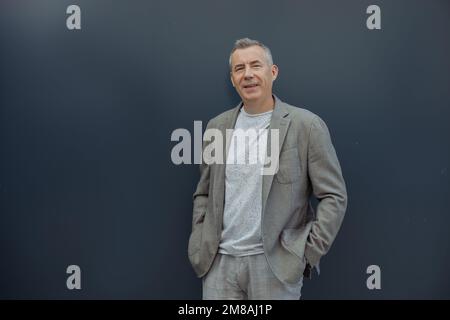 Lächelnder, grinsender, selbstbewusster und cooler erwachsener Mann im Anzug, der mit Ausdruck posiert, mit Händen in Taschen an der blauen Wand Stockfoto