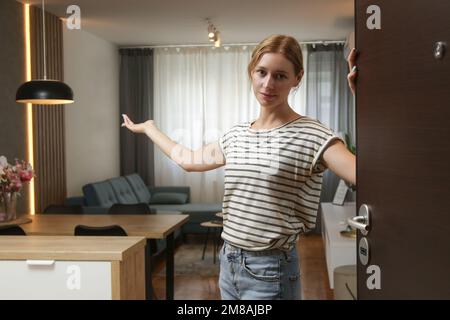 Fröhliche Frau, die die Haustür öffnet und Freunde einlädt. Junge Frau begrüßt Besucher in ihrem Haus. Stockfoto