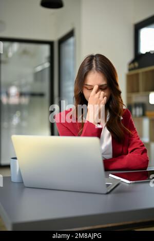 Porträt, gestresst und deprimiert die asiatische Geschäftsfrau der Millennials sitzt an ihrem Schreibtisch, leidet unter Augenbelastung oder Kopfschmerzen und macht sich Sorgen um ihren Profi Stockfoto
