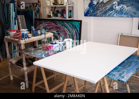 Interieur des Workshops für kreative Kunst. Verschiedene Eimer mit Farbe, Glasgefäße, große weiße Leinwand auf Holzschreibtisch mit Flecken. Stockfoto