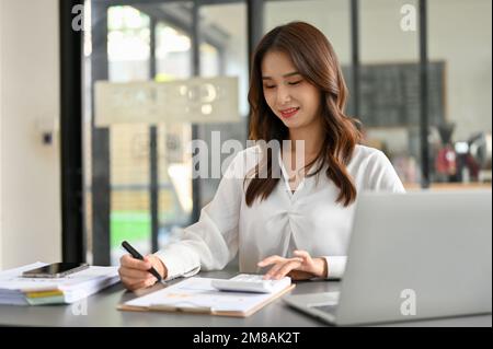 Charmante und fokussierte Buchhalterin der Millennials in Asien, die an ihrem Schreibtisch im Büro arbeitet, Verkaufsberichte verwaltet und einen Taschenrechner verwendet. Stockfoto
