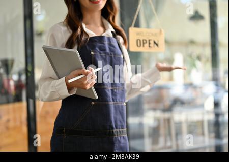 Freundliche junge asiatische Kellnerin oder Café-Angestellte mit ihrem Tablet, steht an der Eingangstür ihres Cafés und begrüßt Gäste. Freigestellte Aufnahme Stockfoto