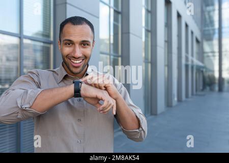 Lateinamerikanischer Geschäftsmann im Hemd, fröhliche Gespräche mit Freunden, die Smartwatch benutzen, Mann außerhalb des Bürogebäudes, der Gadget für Befehle der künstlichen Intelligenz verwendet. Stockfoto