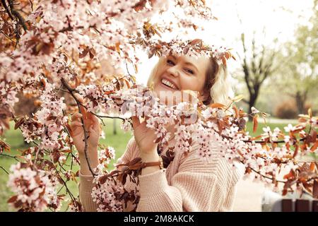 Porträt einer jungen Frau mit pinkfarbener Strickjacke, die neben einem Vogelkirschbaum steht, einen Zweig im Park hält, Spaß hat. Stockfoto