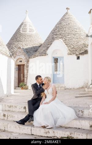 Glückliches Hochzeitspaar, das auf der Treppe in der Nähe von konischen Dächern von Trulli sitzt und sich küsst. Junge Braut umarmt sich mit Bräutigam. Stockfoto