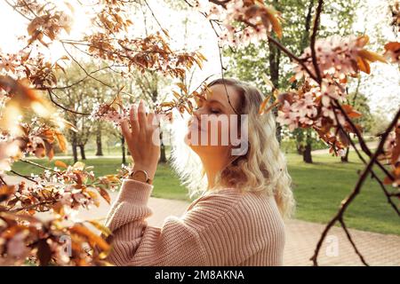 Seitenansicht einer jungen lächelnden Frau, die neben einem blühenden Vogelkirschbaum steht, einen Zweig im Park hält, Blumen schnüffelt. Stockfoto