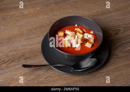 Blick von oben auf die Tomatensuppe mit saurer Sahne und Croutons in schwarzer Schüssel auf Teller mit Löffel auf braunem Holztisch. Stockfoto