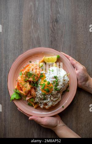 Draufsicht einer unbekannten Frau, die einen großen Keramikteller hält, mit leckeren, sonnigen Eiern, Limette, Lachs, Tomaten. Stockfoto