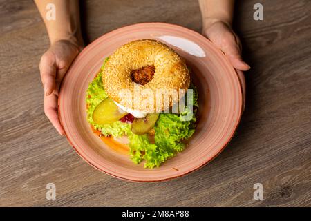 Blick von oben auf eine unbekannte Frau mit leckerem Burger mit Loch im Brötchen, Salat, gebratenem Hähnchen, eingelegten Gurken. Stockfoto