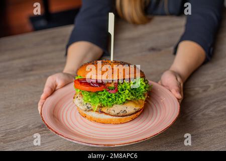 Zugeschnittenes Foto einer Frau, die am Tisch sitzt und einen großen rosa Teller mit frischem Burger hält, mit Salat, Zwiebeln, Tomaten, Hühnchen. Stockfoto