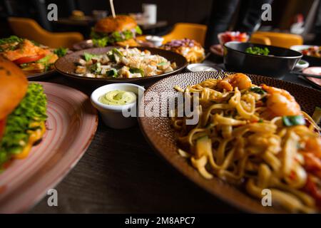 Nahaufnahme verschiedener farbenfroher, appetitlicher Speisen auf einem Holztisch im Restaurant Café. Pasta mit Garnelen. Salat. Burger. Stockfoto