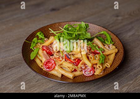Nahaufnahme schmackhafter Pasta mit Kirschtomaten, Rucola-Rucola und Käse, serviert auf brauner, runder Platte auf einem Holztisch. Stockfoto