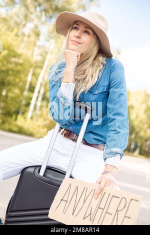 Vertikal positiv lächelnde, unabhängige blonde Frau in Cowgirl hat per Anhalter auf der Straße mit einem Schild überall sitzen auf Gepäck Stockfoto