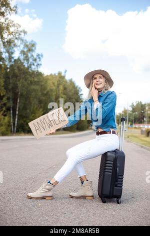 Senkrecht lächelnde, lachende blonde Frau in Cowgirl hat per Anhalter auf der Straße mit Pappteller überall auf dem Koffer sitzen Stockfoto