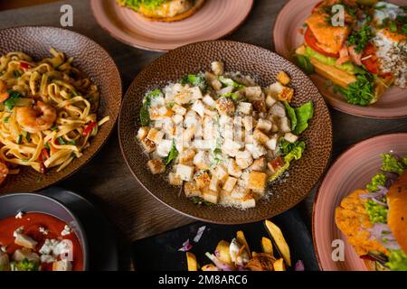 Blick von oben auf Zahnspeisen auf Tellern auf einem Holztisch im Restaurant Cafe. Pasta mit Garnelen. Caesar Salat mit Salat. Stockfoto