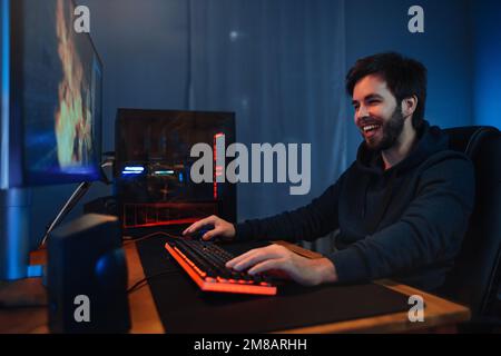 Junger, weißer, glücklicher Gamer, der es genießt, im Online-Shooter zu spielen. Er verbringt seinen Abend zu Hause und spielt Computervideospiele mit Freunden. Cyber-Sport, Computersucht. Seitenansicht, Neonlicht Stockfoto