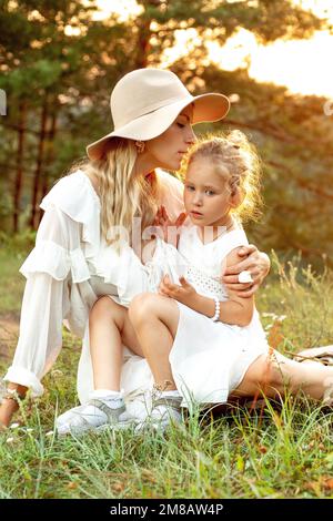 Attraktive Mutter mit kleiner Tochter sitzt in weißen Kleidern auf grünem Gras in Holz bei gelbem Sonnenuntergang. Mama und Kind gehen im Sommer abends im Wald spazieren Stockfoto