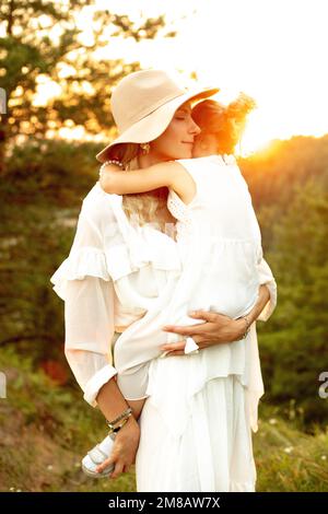 Porträt einer wunderschönen Dame mit Hut und Liebe, die ihr kleines Mädchen bei Sonnenuntergang im Wald in den Händen hält. Mama und Kind gehen im Sommer abends im Wald spazieren Stockfoto