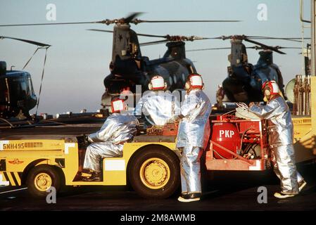 Absturz- und Bergungsmannschaften in Anzügen stehen mit einem MD-3A Feuerwehrfahrzeug während des Flugbetriebs an Bord des Amphibienschiffs USS BELLEAU WOOD (LHA-3) auf dem Cockpit bereit. Marine Medium Helicopter Squadron 161 (HMM-161) CH-46 Sea Knight Helicopters werden im Hintergrund geparkt. Land: Pazifik (POC) Stockfoto