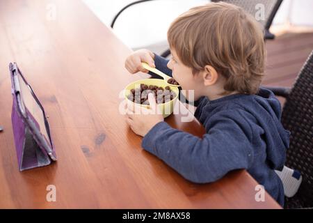 Der kleine Junge sitzt auf dem Stuhl am Tisch und sieht sich mit Interesse den Zeichentrickfilm auf dem Tablet-Computer an. Ein Kind im Kindergartenalter, das den Mund aufmacht, um einen Löffel zu essen Stockfoto