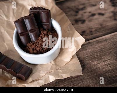 Dunkle poröse Schokoladenstücke und Kakaopulver auf Holzhintergrund. Bitterschokolade, selektiver Fokus Stockfoto