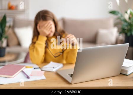 Porträt eines traurigen Mädchens, das müde vom Lernen ist, von zu Hause aus online mit einem Laptop lernt, eine Brille in der Hand hält, erschöpft von Schulaufgaben. Heimunterricht, Fernunterricht durch Covid Stockfoto