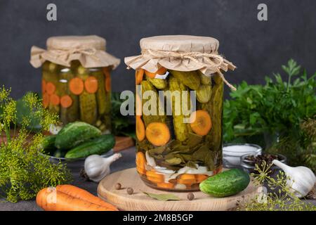 Selbstgemachte eingelegte Gurken mit Karotten, Knoblauch, Lorbeerblättern und Dill in zwei Gläsern auf grauem Hintergrund Stockfoto