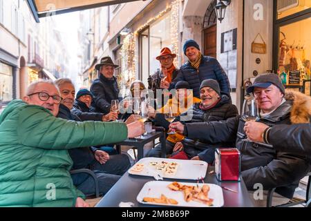 Eine Gruppe erwachsener Männer feierte im Winter mit Freunden im Ruhestand einen Geburtstag, indem sie an den Tischen eines Cafés im Freien anstoßen Stockfoto