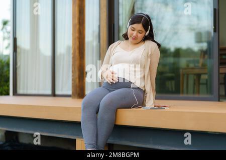 Porträt einer schwangeren Happy Asian Woman, die zu Hause auf der Terrasse sitzt und über ein Smartphone Musik über Kopfhörer hört. Stockfoto