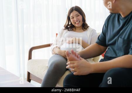 Der asiatische Ehemann massiert die Füße der schwangeren Frau im Wohnzimmer des Hauses. Glückliche schwangere Familie. Stockfoto