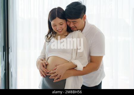 Glückliches asiatisches Paar erwartet ein Baby, das zu Hause am Fenster steht, liebevoller Ehemann, der zärtlich den Bauch seiner schwangeren Frau berührt Stockfoto