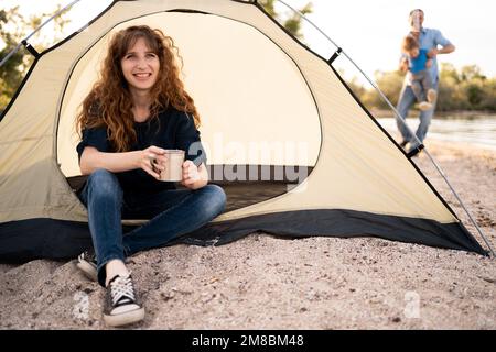 Eine junge Frau trifft auf einen kalten Morgen in einem touristischen Campingzelt mit einer Tasse heißem Tee oder Kaffee. Romantisches Campingkonzept. Stockfoto