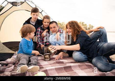 Glückliche Familie ruht in einem Zelt in der Natur. Sommerurlaub mit Kindern. Große Familie mit drei Kindern, die draußen Tee auf dem Campingplatz trinken. Verstanden Stockfoto