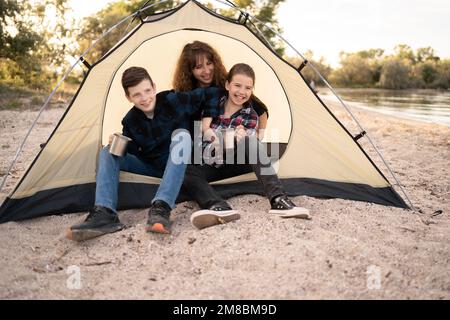 Porträt einer Familie von Reisenden im Zelt, die in die Kamera schauen. Lächelnde Mutter, Mädchen und Junge, die vor dem Zelt glücklich aussehen. Natur auf Familienautou erkunden Stockfoto
