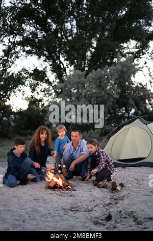 Glückliche große Familie, die am Abend Marshmallow über dem Lagerfeuer röstet. Camping, Reisen, Tourismus, Wandern und Menschen Konzept Stockfoto