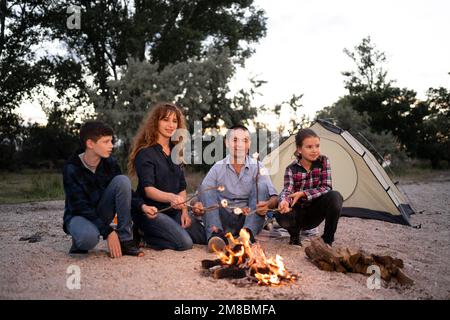 Eine vierköpfige Familie, Dad, mom, Sohn und Tochter, sitzen am Feuer. Warme Marshmallows und Erholung im Freien. Urlaubskonzept in t Stockfoto