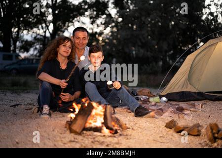 Familie - Mutter, Vater und Sohn rösten Marshmallow am Lagerfeuer. Reisen, Tourismus, Wandern, Picknick und Menschenkonzept Stockfoto