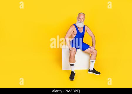 Großes Porträt eines extravaganten, sportlichen alten Menschen, der auf dem Podium sitzt, leerer Platz isoliert auf gelbem Hintergrund Stockfoto