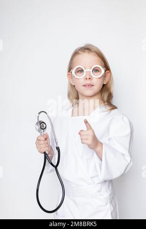 Ernstes kleines Mädchen in Arztkleidung mit Spielzeugbrille, Stethoskop in der Hand, Zeigefinger nach oben in Stopp- oder Verbotsbewegung. Ge Stockfoto