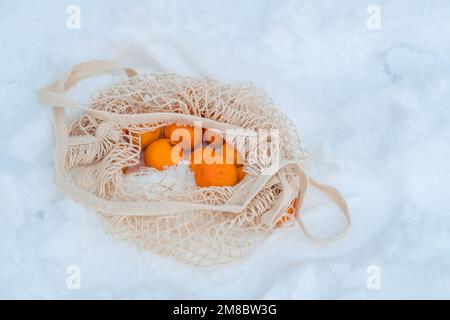 Die Mandarinen sind in einem Sack aus organischem Gewebe auf einer Bank, die mit Schnee bedeckt ist. Winterferienkonzept Stockfoto