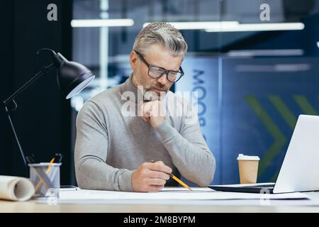 Nahaufnahme. Konzentrierter leitender, grauhaariger Architekt, Ingenieur, Designer, der mit einem Laptop im Büro sitzt und mit drawings.plans arbeitet. Macht Notizen, Papierkram. Stockfoto